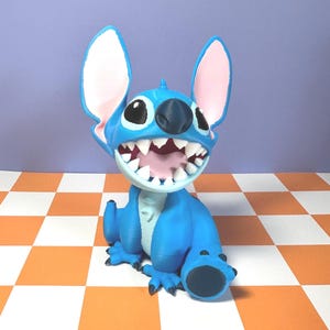 Puede incluir: Figura azul de Stitch de la pel&iacute;cula Lilo & Stitch. La figura tiene orejas grandes con el interior rosa, una boca abierta con dientes blancos y est&aacute; sobre una superficie a cuadros naranja y blanco. Personaje popular.