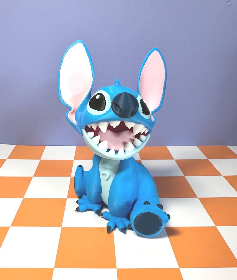 Puede incluir: Figura azul de Stitch de la pel&iacute;cula Lilo & Stitch. La figura tiene orejas grandes con el interior rosa, una boca abierta con dientes blancos y est&aacute; sobre una superficie a cuadros naranja y blanco. Personaje popular.
