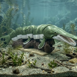 May include: A segmented, articulated fish figurine with a long, slender snout and an open mouth showing teeth. The body is olive green with dark spots on the top and white on the underside, featuring visible joints between sections, resting on a base.