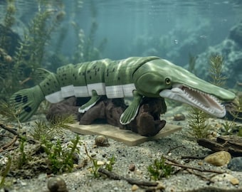 May include: A segmented, articulated fish figurine with a long, slender snout and an open mouth showing teeth. The body is olive green with dark spots on the top and white on the underside, featuring visible joints between sections, resting on a base.