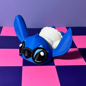 May include: A blue 3D-printed figure of a cartoon character with large ears, black eyes, and a white plush interior. The figure is lying down on a pink and blue checkered surface.