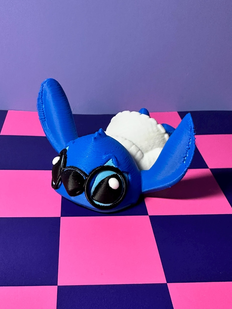 May include: A blue 3D-printed figure of a cartoon character with large ears, black eyes, and a white plush interior. The figure is lying down on a pink and blue checkered surface.