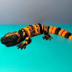 May include: A detailed, three-dimensional model of a Gila monster lizard. The lizard features a black head and limbs, with a segmented body in alternating bands of black and bright orange. The model is resting on a turquoise surface.
