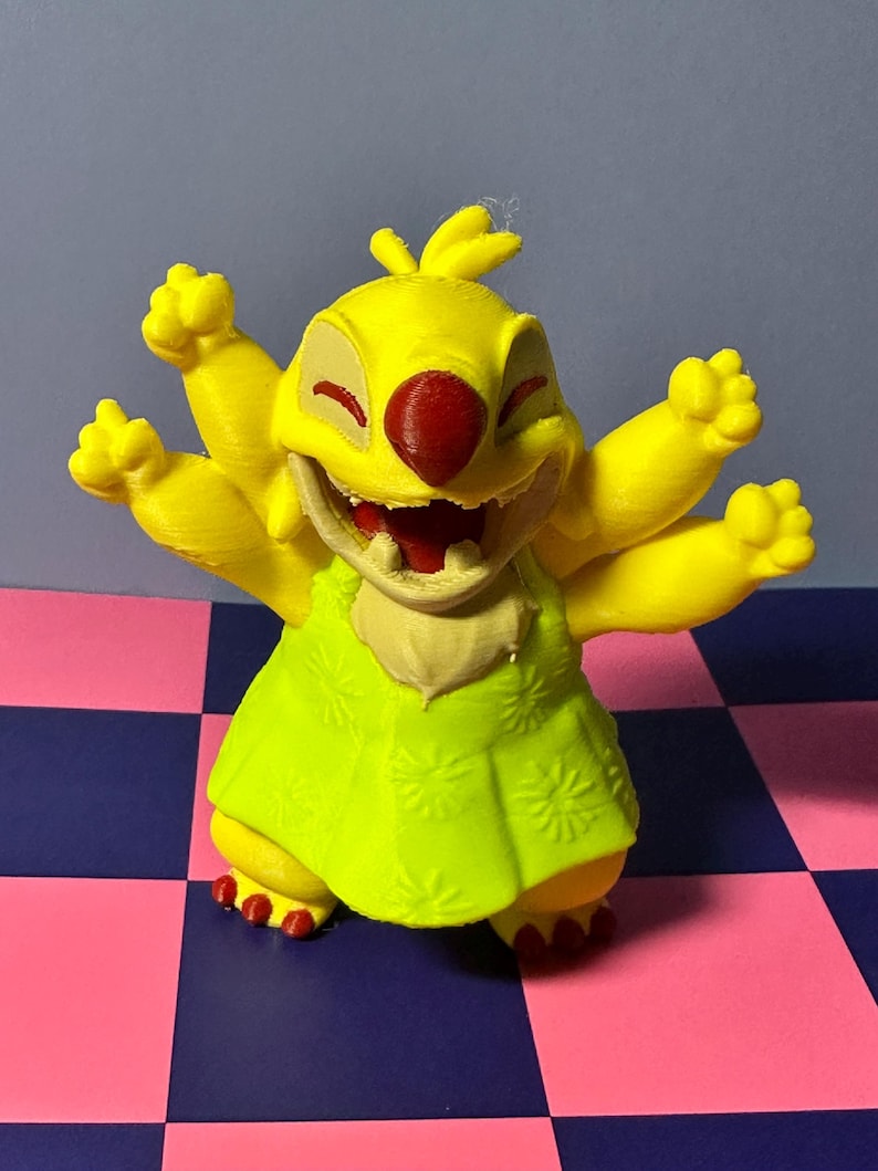 May include: A yellow figurine of a cartoon character with six arms, a red nose, and a green dress. The figurine is smiling with its mouth open. The background is a checkered pattern of pink and blue squares.