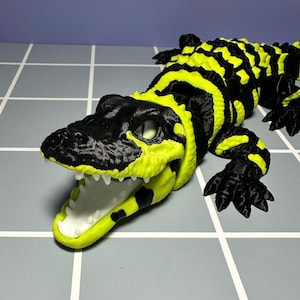 May include: A 3D-printed alligator toy in black and neon yellow. The alligator has an open mouth with white teeth and a detailed textured body. The toy is lying on a gray surface with white lines.
