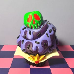 May include: A decorative cupcake with purple frosting, black sprinkles, and a green skull with red accents on top. The cupcake sits on a white square with a gold and red crown at the base. The background is a checkered pattern.