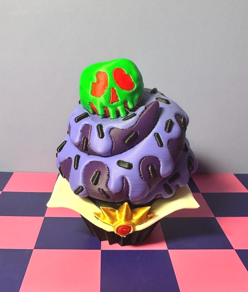 May include: A decorative cupcake with purple frosting, black sprinkles, and a green skull with red accents on top. The cupcake sits on a white square with a gold and red crown at the base. The background is a checkered pattern.