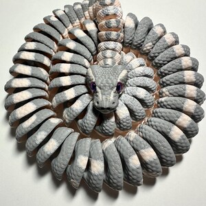 Puede incluir: Escultura en espiral de una serpiente, con un patr&oacute;n gris y blanco. La cabeza de la serpiente est&aacute; centrada, con ojos morados. El cuerpo est&aacute; hecho de segmentos curvos, creando una forma circular. La escultura est&aacute; sobre un fondo blanco.