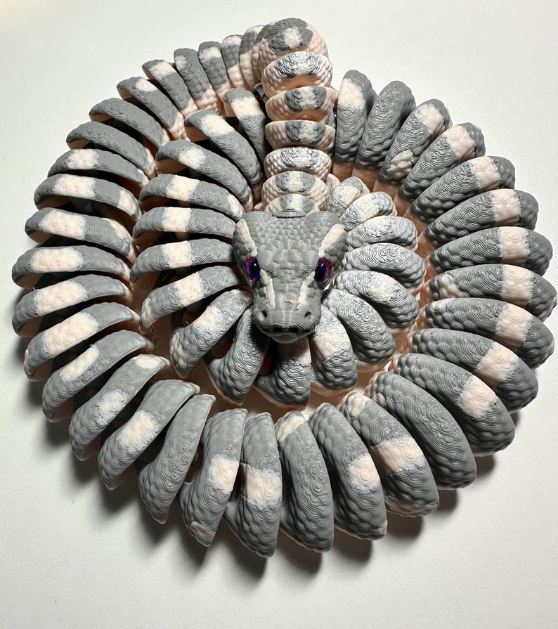 Puede incluir: Escultura en espiral de una serpiente, con un patr&oacute;n gris y blanco. La cabeza de la serpiente est&aacute; centrada, con ojos morados. El cuerpo est&aacute; hecho de segmentos curvos, creando una forma circular. La escultura est&aacute; sobre un fondo blanco.