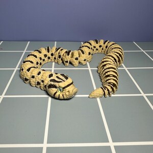 May include: A beige and black toy snake with a detailed design. The snake has a segmented body with black spots and stripes. The head features green eyes and a detailed, textured surface. The toy is posed on a gray surface with a grid pattern.