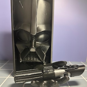 May include: A black 3D-printed Darth Vader bust with a lightsaber. The bust is rectangular and features the iconic helmet and face of Darth Vader. The lightsaber is held in a hand-shaped grip, with a silver and black design.