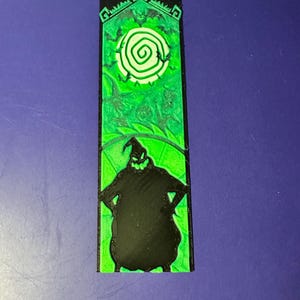 Puede incluir: Un objeto decorativo rectangular verde y negro que presenta la silueta de un personaje con los brazos en jarras. La parte superior tiene un dise&ntilde;o verde con una espiral blanca. El objeto est&aacute; sobre un fondo azul.
