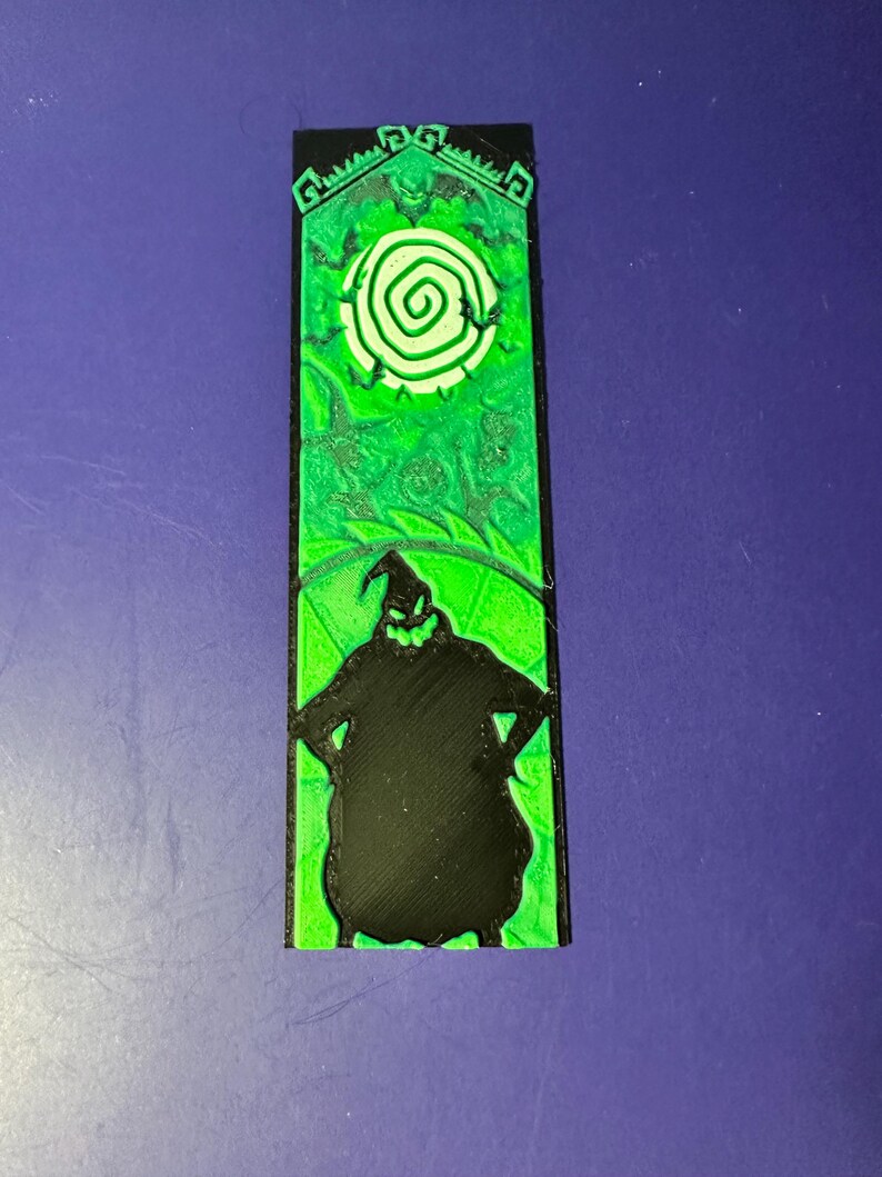 Puede incluir: Un objeto decorativo rectangular verde y negro que presenta la silueta de un personaje con los brazos en jarras. La parte superior tiene un dise&ntilde;o verde con una espiral blanca. El objeto est&aacute; sobre un fondo azul.