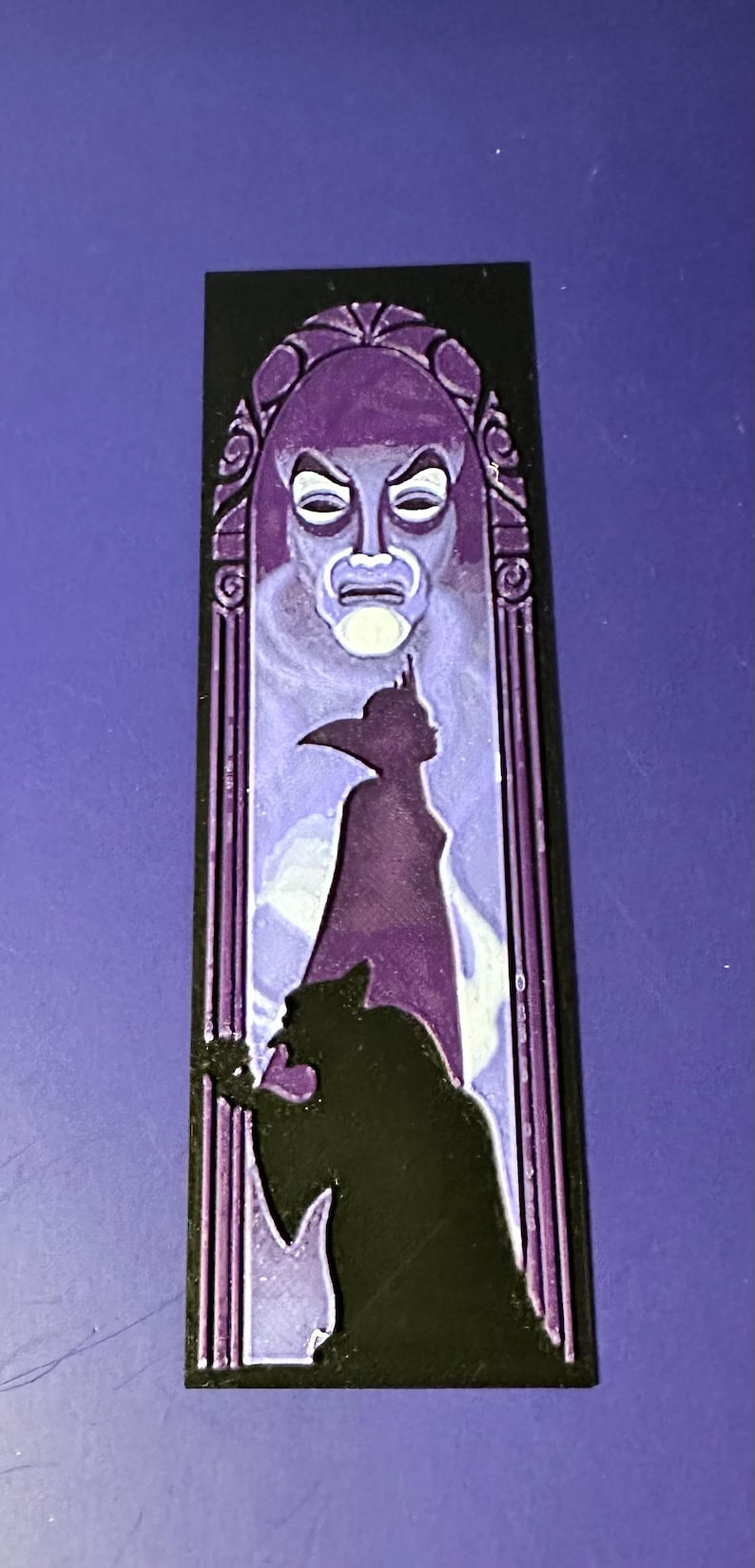 Puede incluir: Una pieza decorativa rectangular negra y morada con una imagen estilizada de un personaje de cuento de hadas. El dise&ntilde;o incluye una cara, una silueta de una figura y una silueta oscura.