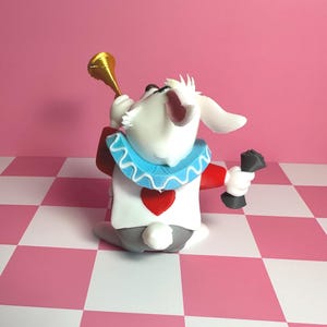 Pode incluir: Uma figura extravagante do Coelho Branco de Alice no Pa&iacute;s das Maravilhas. O coelho &eacute; branco com uma roupa vermelha e branca, uma gola azul com babados e uma trombeta dourada. Ele segura um objeto preto na m&atilde;o direita, em um fundo xadrez rosa e branco.