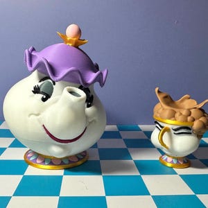 May include: A white teapot with a purple lid and a smiling face, next to a matching teacup. The teapot has a pink accent on top and a gold and pink base. The teacup has a gold handle and a brown top.