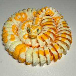 Puede incluir: Figura de serpiente enroscada, impresa en 3D, en tonos amarillo y crema. El cuerpo de la serpiente est&aacute; segmentado con un patr&oacute;n repetitivo de manchas amarillas. La cabeza de la serpiente est&aacute; detallada con ojos verdes y un patr&oacute;n amarillo y crema.