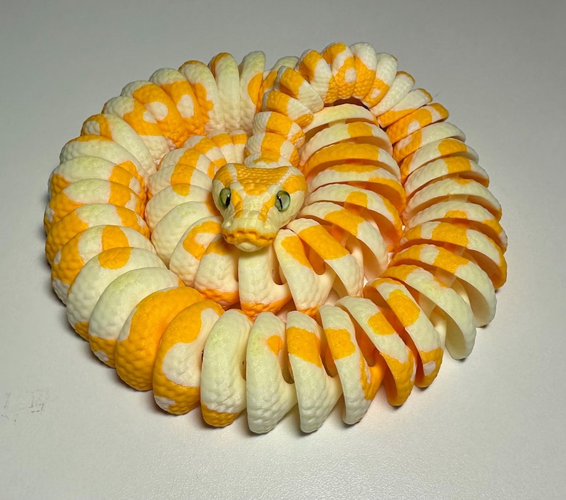Puede incluir: Figura de serpiente enroscada, impresa en 3D, en tonos amarillo y crema. El cuerpo de la serpiente est&aacute; segmentado con un patr&oacute;n repetitivo de manchas amarillas. La cabeza de la serpiente est&aacute; detallada con ojos verdes y un patr&oacute;n amarillo y crema.