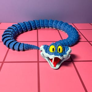 May include: A blue articulated snake toy with a white and blue head, yellow eyes, and a red tongue. The toy is posed on a pink tiled surface with a purple background. The snake's body is segmented.