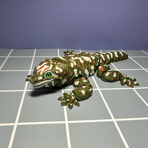 May include: A detailed, green and white spotted gecko figurine with red accents. The reptile has bright green eyes and is lying on a gray tiled surface. The gecko's body is textured, and its feet are detailed.