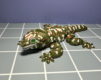 May include: A detailed, green and white spotted gecko figurine with red accents. The reptile has bright green eyes and is lying on a gray tiled surface. The gecko's body is textured, and its feet are detailed.