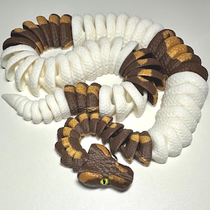 Puede incluir: Un juguete de serpiente articulada impreso en 3D en blanco, marr&oacute;n y amarillo. El cuerpo de la serpiente est&aacute; segmentado, lo que permite un movimiento flexible. La cabeza presenta escamas detalladas y un ojo amarillo. El juguete est&aacute; dise&ntilde;ado para parecerse a una serpiente realista.