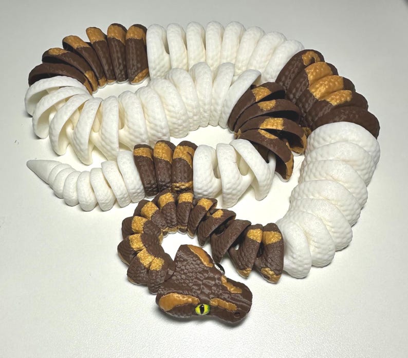 Puede incluir: Un juguete de serpiente articulada impreso en 3D en blanco, marr&oacute;n y amarillo. El cuerpo de la serpiente est&aacute; segmentado, lo que permite un movimiento flexible. La cabeza presenta escamas detalladas y un ojo amarillo. El juguete est&aacute; dise&ntilde;ado para parecerse a una serpiente realista.