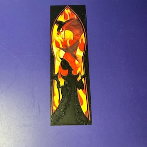 Puede incluir: Un marcap&aacute;ginas rectangular negro con un dise&ntilde;o de estilo vidriera. El dise&ntilde;o incluye un drag&oacute;n rojo en llamas, con la silueta de una figura sosteniendo un bast&oacute;n. El fondo es amarillo y naranja.
