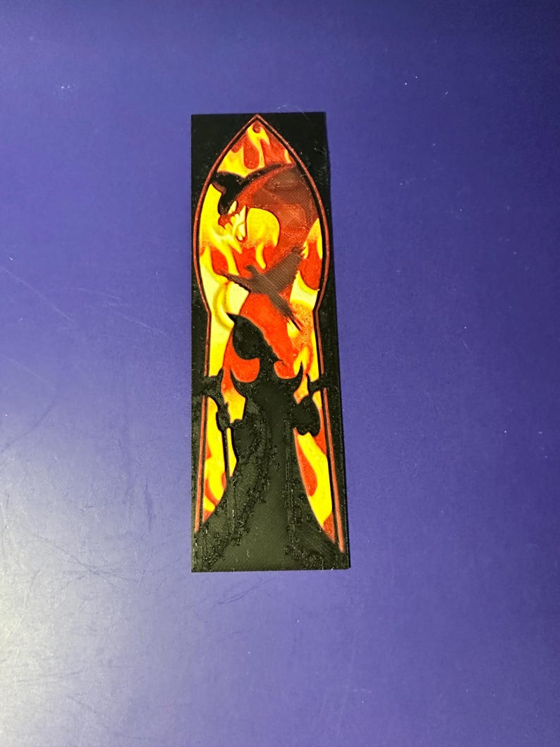 Puede incluir: Un marcap&aacute;ginas rectangular negro con un dise&ntilde;o de estilo vidriera. El dise&ntilde;o incluye un drag&oacute;n rojo en llamas, con la silueta de una figura sosteniendo un bast&oacute;n. El fondo es amarillo y naranja.