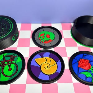 May include: A collection of round coasters with black rims and colorful designs. The designs include a green skull, a red rose, a yellow seashell, and a green and red bicycle. A black container with a green rim is also present.