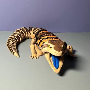 May include: A detailed, articulated lizard toy with a segmented body in shades of brown, black, and orange. The lizard has a tan head with yellow eyes and a blue tongue. The tail is curled and the legs are extended.