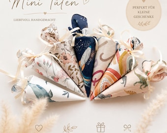 May include: A collection of seven cone-shaped fabric gift bags tied with cream ribbons. The bags feature various patterns including floral prints, a dark blue space theme with a rocket, rainbow motifs, and dinosaur illustrations on light backgrounds.