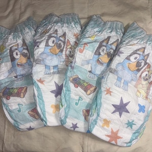 May include: Four disposable diapers featuring a cartoon dog character in blue, with musical notes, stars, and a xylophone design. The diapers have a light blue and white color scheme.