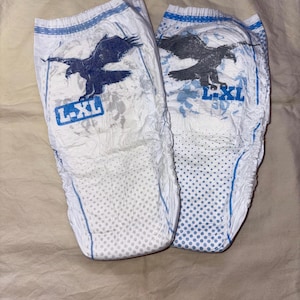 Puede incluir: Dos pañales desechables blancos con un gráfico de águila azul y el texto "L-XL". Los pañales tienen un ribete azul y un patrón de puntos en la parte delantera. Los pañales están diseñados para bebés y niños pequeños.