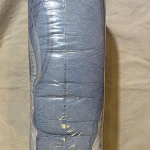 Może przedstawiać: Opakowanie jasnoniebieskich pieluch dla dorosłych, zapieczętowane w przezroczystym plastiku. Pieluchy są złożone i ułożone w stos, z widocznym szwem pośrodku. Opakowanie jest lekko pomarszczone.