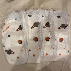 May include: Four white baby diapers with an outer space theme. Each diaper features a repeating pattern of planets, stars, and shooting stars in shades of brown, orange, and green. The diapers are folded and ready for use.