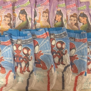 Op de afbeelding: Een verzameling baby luiers met Disney prinsessen en Spider-Man. De luiers hebben kleurrijke illustraties van Jasmine, Belle, Mulan en Spider-Man. De luiers zijn blauw en paars met rode en witte accenten.