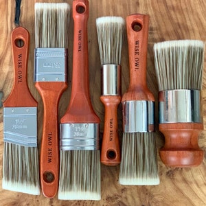 May include: Set of six paint brushes with wooden handles and silver metal ferrules. The brushes have different sizes and shapes, including round and flat. The handles are engraved with the text "WISE OWL".