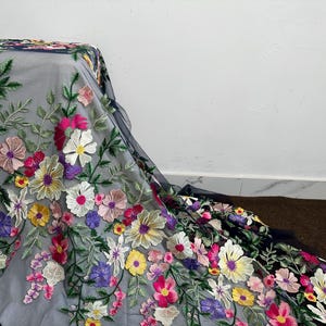 May include: Embroidered floral fabric with a variety of colourful flowers in shades of pink, white, yellow, and purple. The flowers are set against a grey background with green leaves. The fabric is draped over a surface.