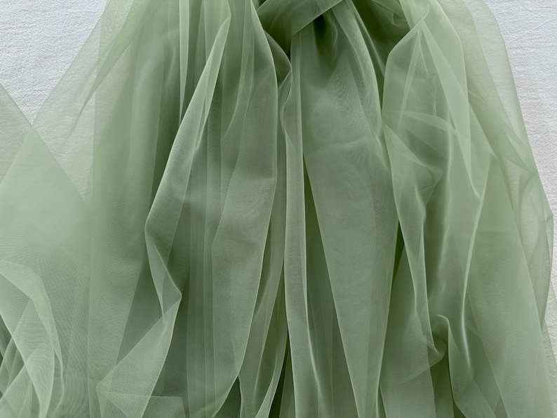May include: A piece of sage green tulle fabric, draped and folded, showing the sheer, delicate texture of the material.