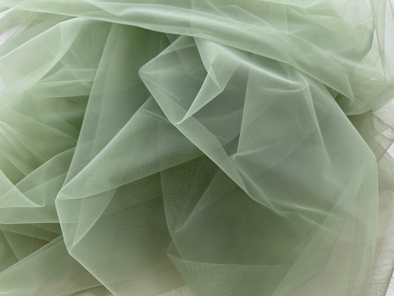 May include: A close-up of a sage green tulle fabric. The fabric is soft and sheer, with a delicate texture.