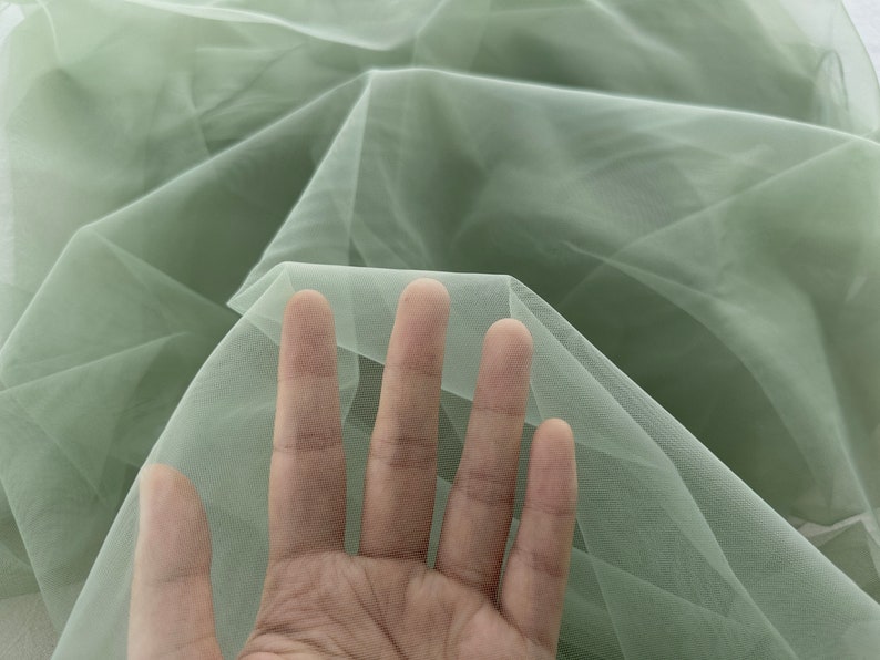 May include: A piece of soft, sage green tulle fabric. The fabric is sheer and delicate, with a fine mesh weave.
