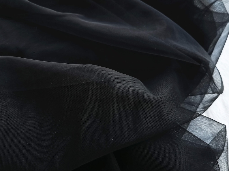 May include: Close-up of black tulle fabric with a soft, flowing texture. The fabric is gathered and draped, creating a delicate and airy look.