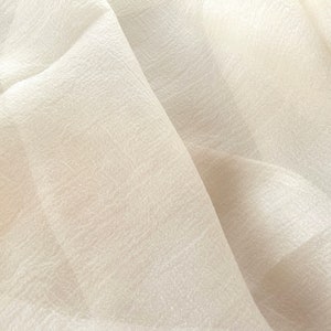 Puede incluir: Primer plano de una tela blanca transparente con una superficie texturizada. La tela est&aacute; doblada y drapeada, creando pliegues y arrugas suaves.