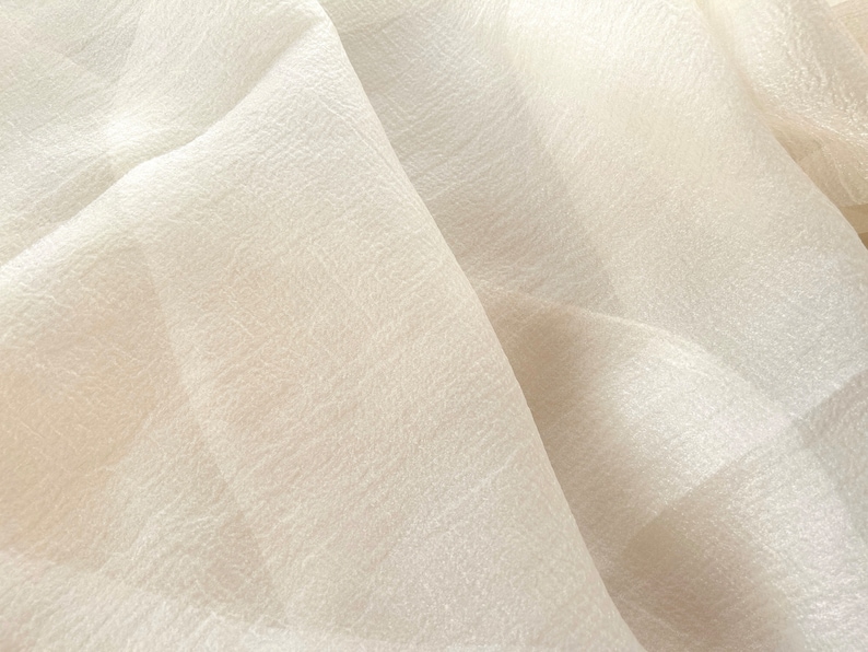 Puede incluir: Primer plano de una tela blanca transparente con una superficie texturizada. La tela est&aacute; doblada y drapeada, creando pliegues y arrugas suaves.