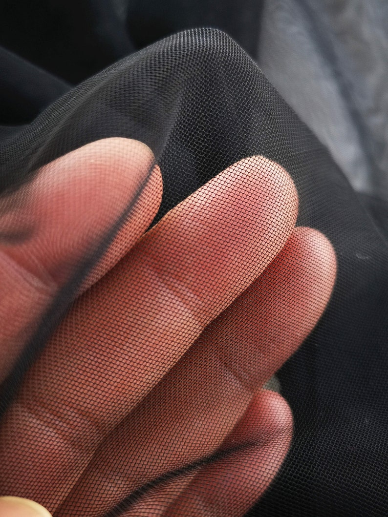 May include: Black sheer fabric with a fine mesh pattern. The fabric is held in a hand, showing the texture and transparency.