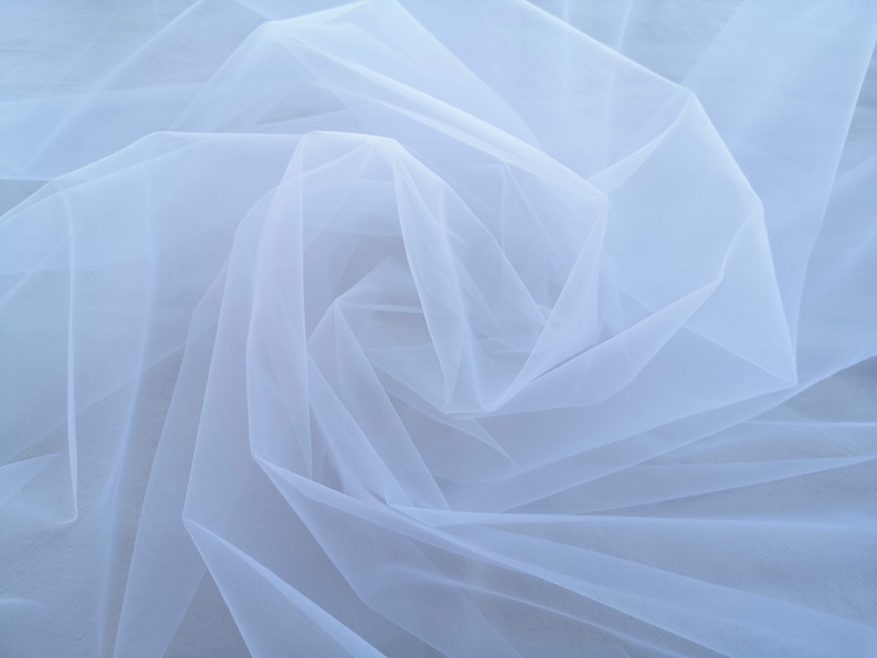 May include: White tulle fabric with a soft, delicate texture. The fabric is draped and folded, creating a flowing and ethereal look.
