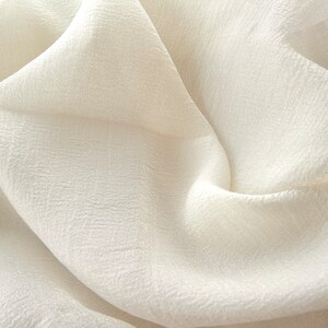 Puede incluir: Primer plano de una tela blanca con superficie texturizada. La tela est&aacute; doblada y drapeada, creando pliegues y arrugas suaves.