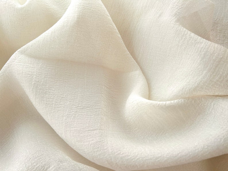 Puede incluir: Primer plano de una tela blanca con superficie texturizada. La tela est&aacute; doblada y drapeada, creando pliegues y arrugas suaves.