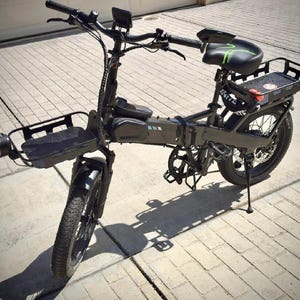 May include: A black electric bicycle with a front basket, rear cargo rack, and a black seat with green accents. The bike has a headlight, fenders, and a kickstand. The tires have a thick tread.